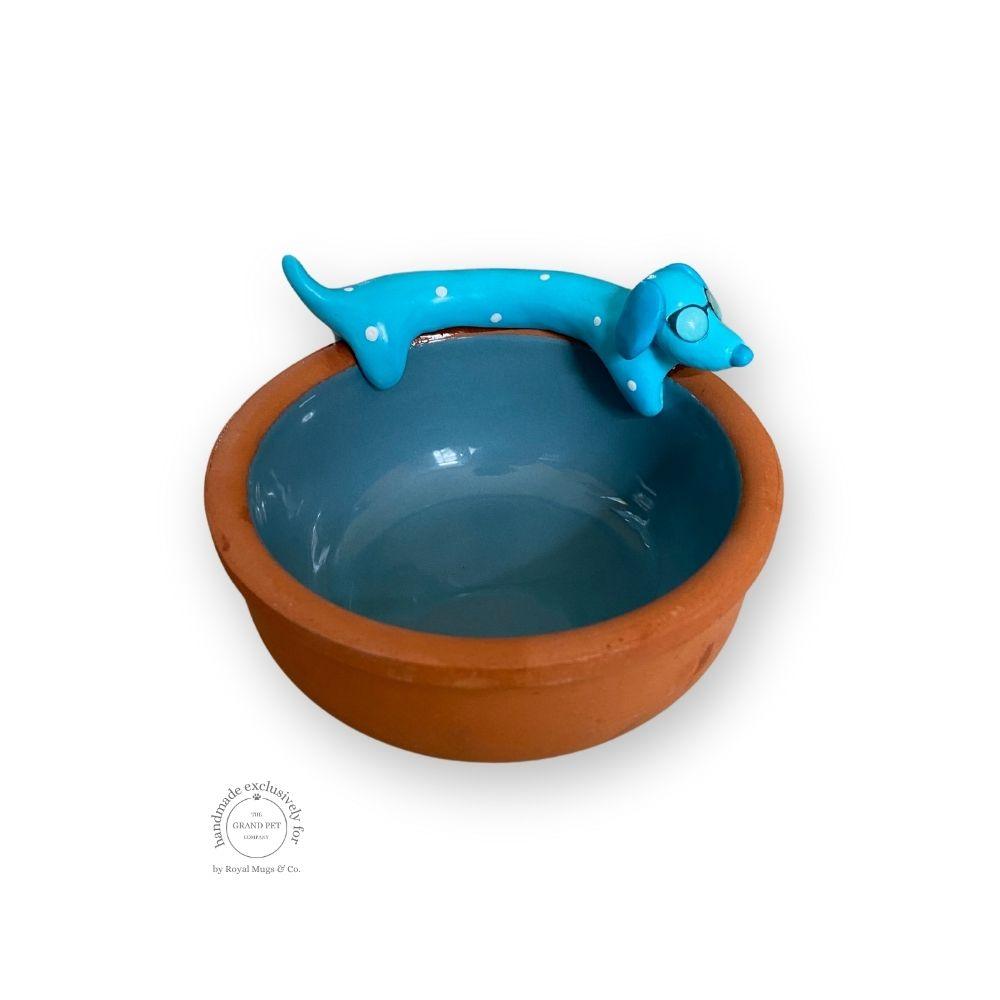A ceramic terracotta pet feeding bowl with a blue interior and a hand-sculpted blue dachshund figure on the side.