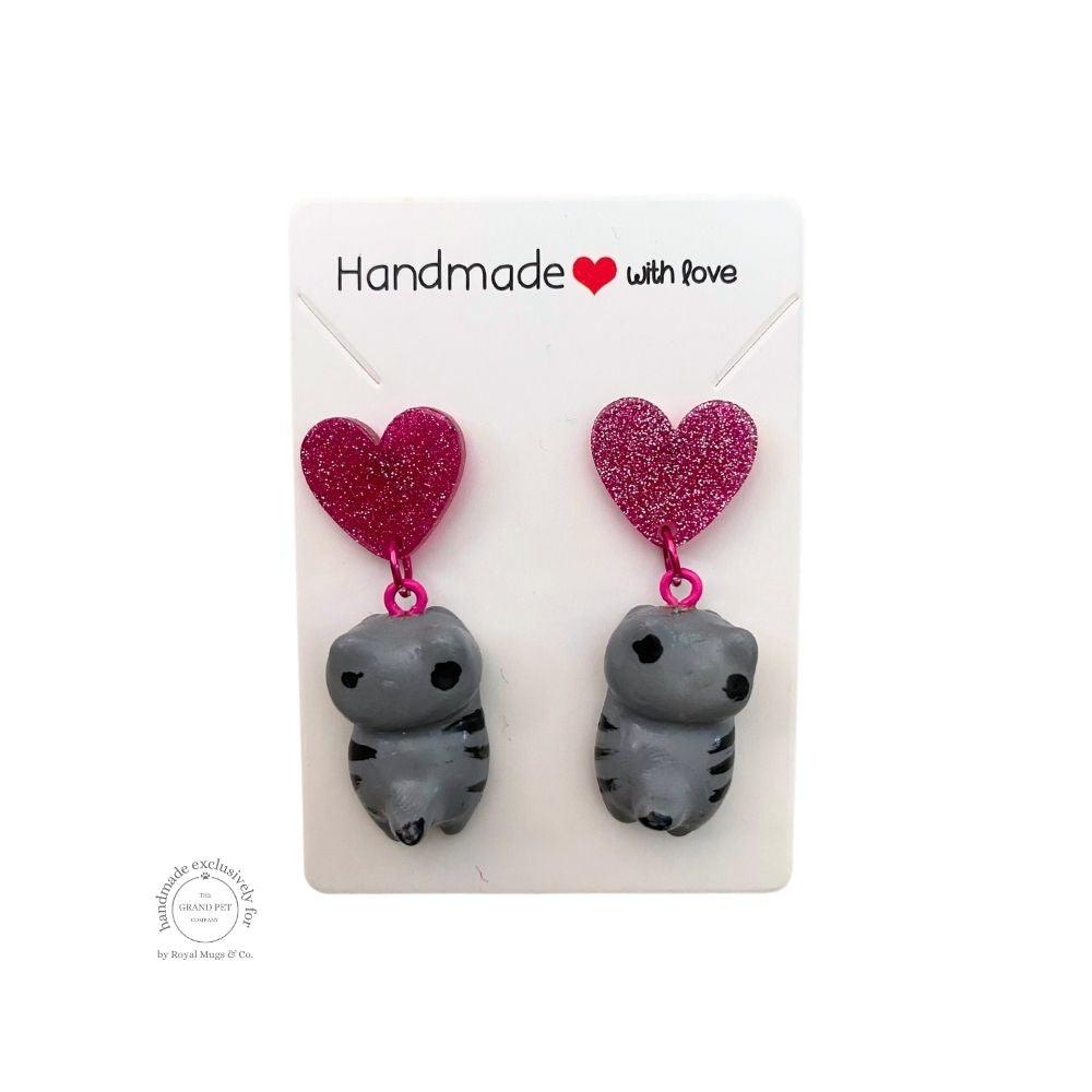 A pair of handcrafted earrings made with polymer clay, featuring a grey cat and a dark pink glitter heart on a clear backing card with 'HANDMADE WITH LOVE' text.