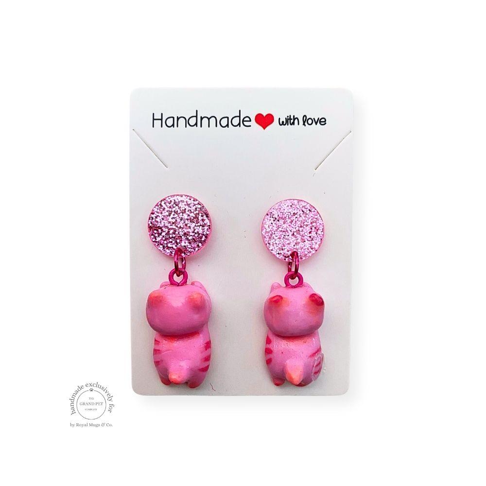 A pair of pink polymer clay earrings designed in the shape of cat with round pink glitter studs, packaged on a white backing card with 'Handmade with love' text.