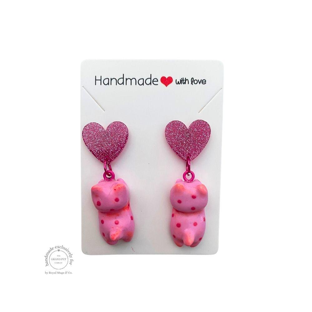 A pair of pink polymer clay earrings in the shape of cats with pink hearts hanging off a dark pink glitter heart, displayed on a white backing card with 'handmade with love' text.