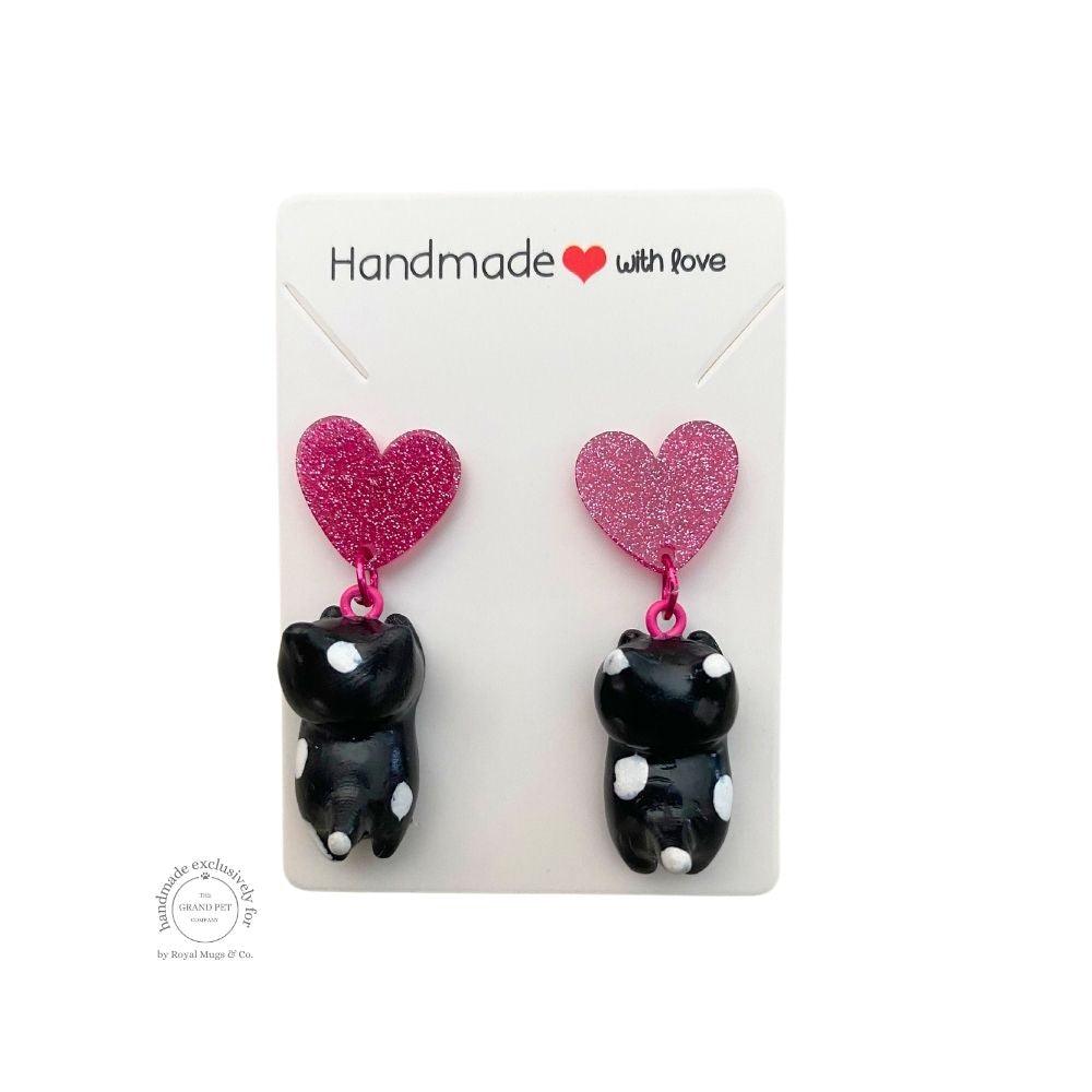 A pair of handcrafted polymer clay earrings in the shape of a cat with a dark pink glitter heart attached to the top, packaged on a white backing card with 'HANDMADE WITH LOVE' text.