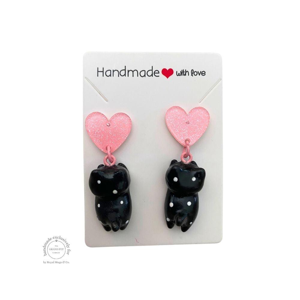A pair of handcrafted polymer clay earrings in the shape of a black and white polka cat with a pink glitter heart attached to the top, packaged on a white backing card with 'HANDMADE WITH LOVE' text.