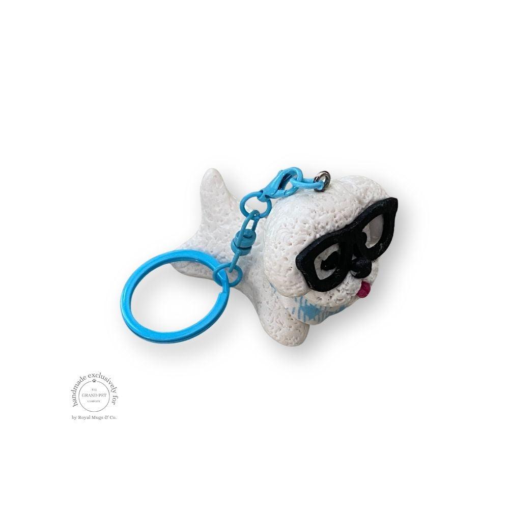 A hand-sculpted blue keychain depicting a white poodle with black glasses and a bandana.
