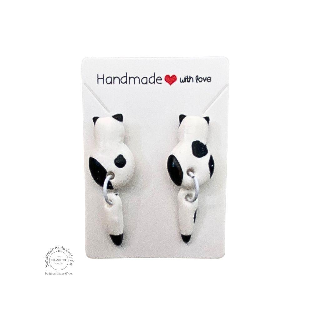 A pair of white and black cat-shaped earrings with tails, displayed on a white backing card with the text 'HANDMADE WITH LOVE'.