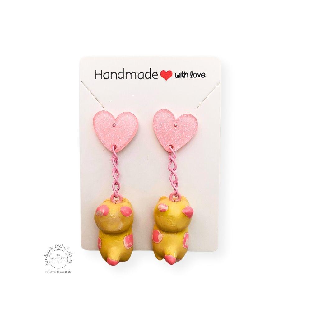 Handmade polymer clay earrings in the shape of a yellow cat with pink glitter hearts, displayed on a white card with 'Handmade with love' text.