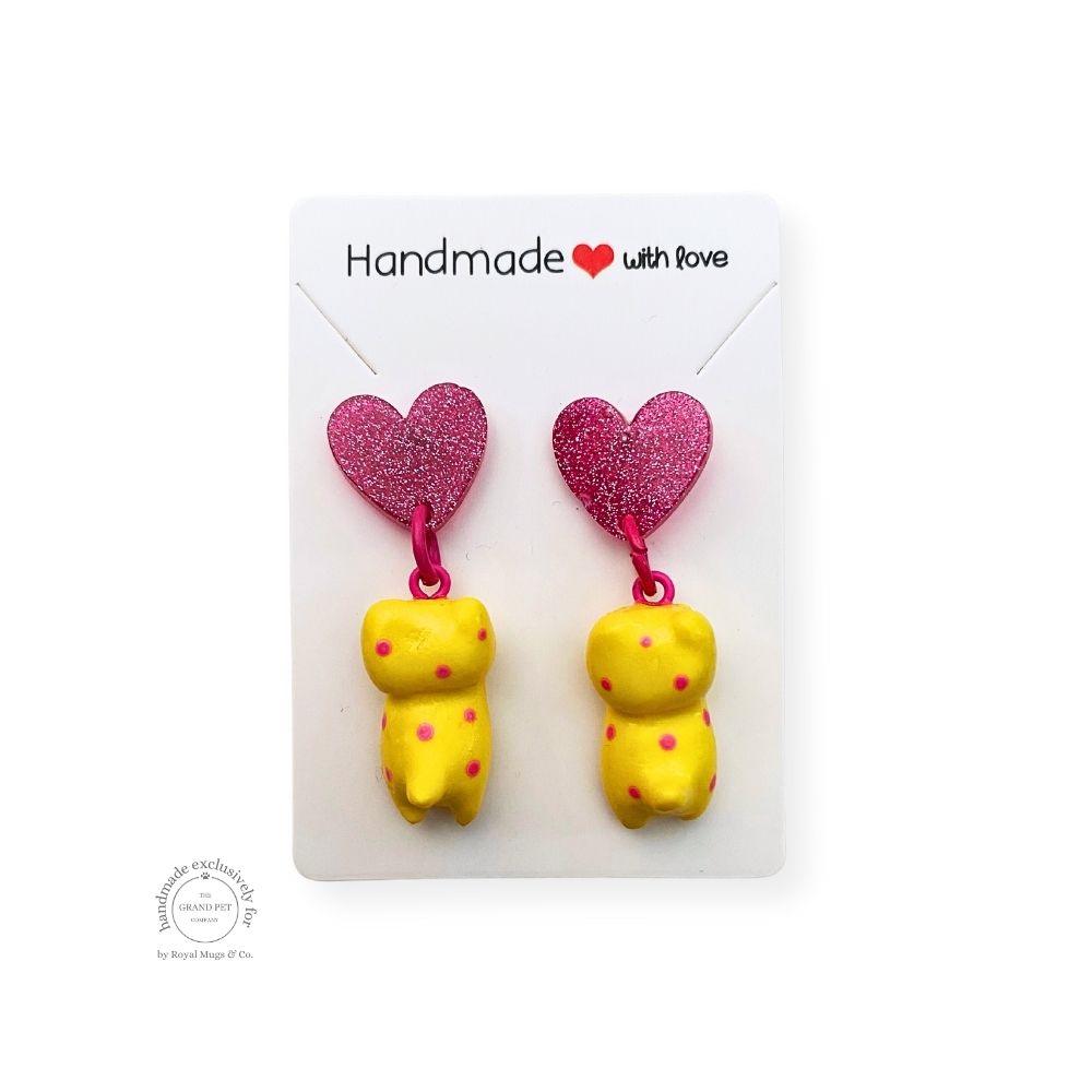 A pair of handcrafted polymer clay earrings in the shape of a yellow and red polka cat with a pink glitter heart attached to the top, packaged on a white backing card with 'HANDMADE WITH LOVE' text.