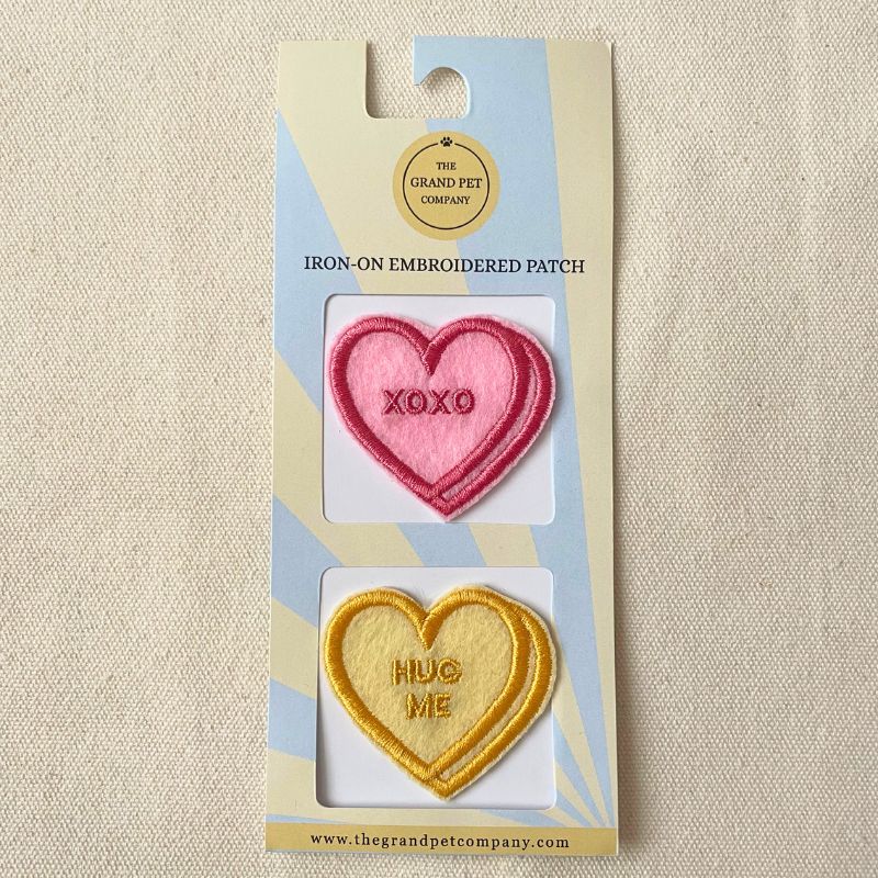 Two Heart-shaped iron-on embroidered felt patches in pink and yellow with text on a Grand Pet Company packaging.
