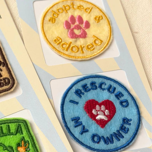 Close-up of embroidered pet adoption patches on cards. One round patch is yellow with a pink paw print saying "ADOPTED & ADORED' and the other is a blue patch with a red heart and white pawprint saying 'I RESCUED MY OWNER'.