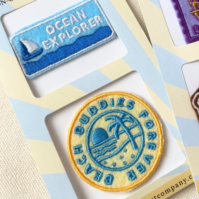 Two embroidered patches displayed on a light blue and cream card: a rectangular blue patch reading "OCEAN EXPLORER" with a sailboat, and a round yellow-and-blue patch reading "BEACH BUDDIES FOREVER" with a beach scene. 