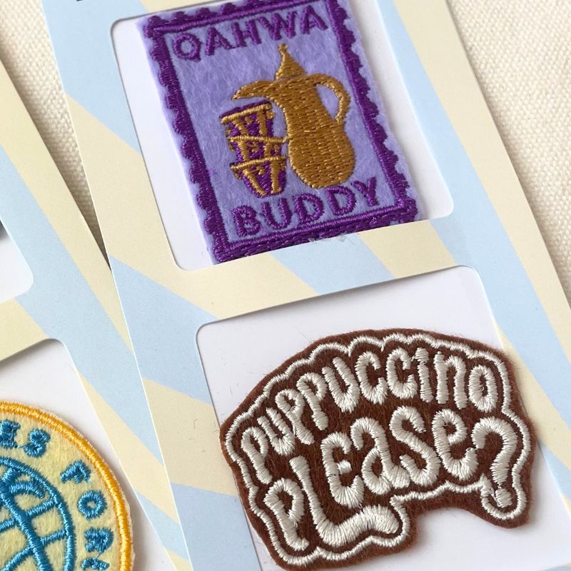 Close-up of embroidered patches mounted on a light blue and cream display card. One patch reads "QAHWA BUDDY" in purple with a gold pitcher motif, and another reads "puppuccino please?" in brown and cream bubble font.
