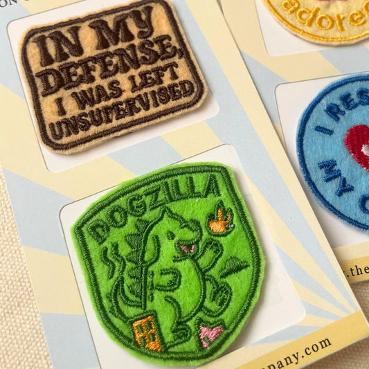 A close-up of embroidered patches displayed on a card. One patch is green with a cartoon dog and the word 'DOGZILLA,' while another patch reads 'IN MY DEFENSE, I WAS LEFT UNSUPERVISED' in brown on a beige background.