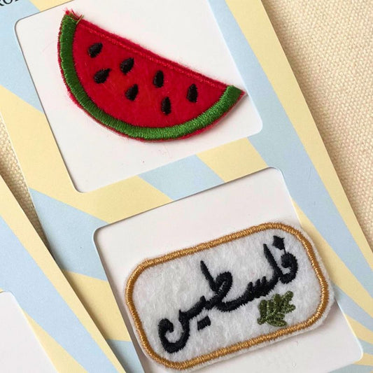 Two embroidered patches displayed on white cards over a pastel striped background. The top patch shows a watermelon slice with red flesh, green rind, and black seeds. The bottom patch says 'Filistine' in Arabic with a green olive branch underneath.