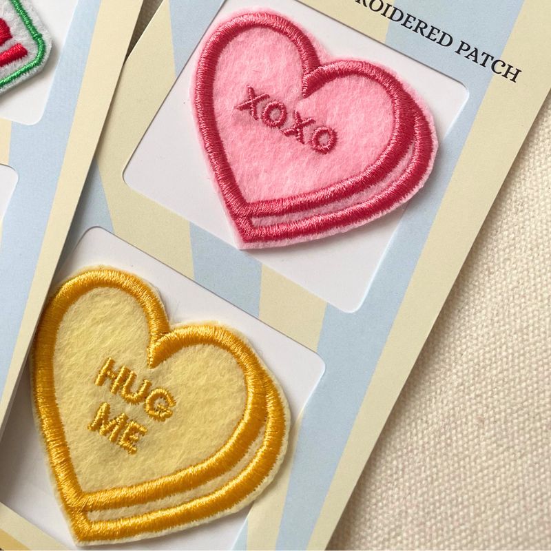 Two embroidered heart-shaped patches on a card, one pink with 'XOXO' in darker pink and one yellow with 'HUG ME' in gold, shown on a beige fabric background.