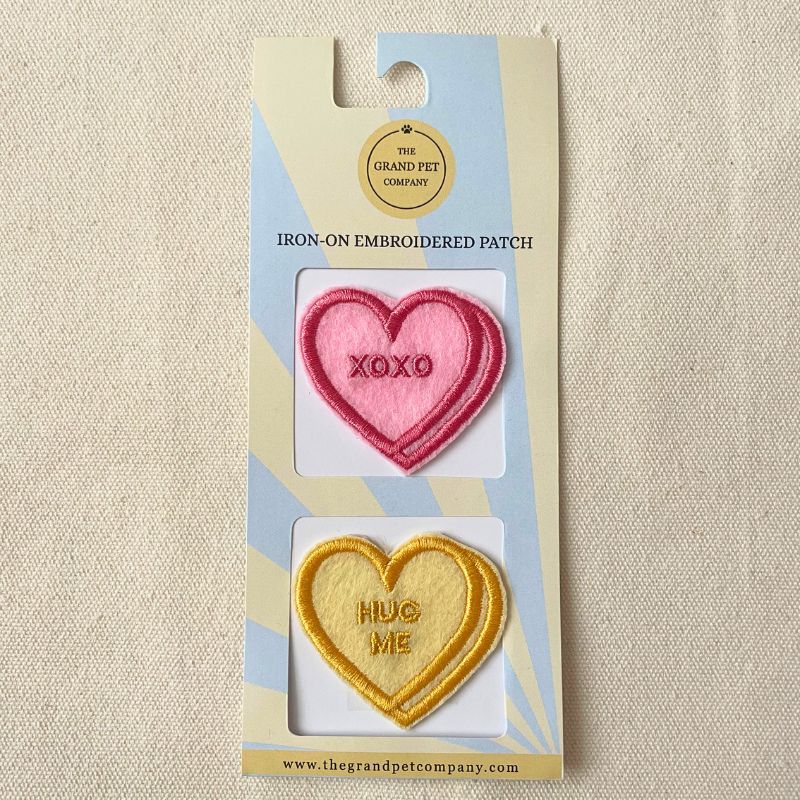 Two Heart-shaped iron-on embroidered felt patches in pink and yellow with text on a Grand Pet Company packaging.