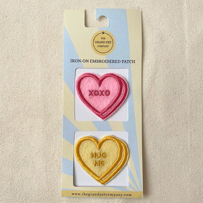 Two Heart-shaped iron-on embroidered felt patches in pink and yellow with text on a Grand Pet Company packaging.