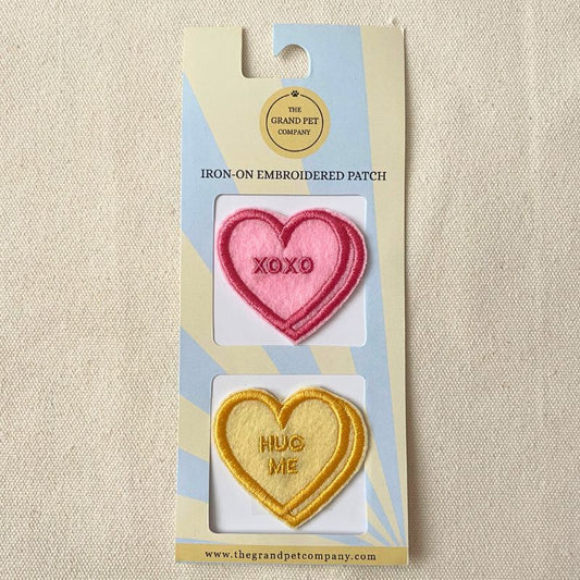 Two Heart-shaped iron-on embroidered felt patches in pink and yellow with text on a Grand Pet Company packaging.