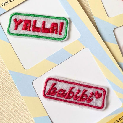 Two embroidered rectangular patches on white backing cards. The top patch reads “YALLA!” in red embroidery with a green border. The second patch is a red-bordered patch reading “HABIBTI” in red on a white background.