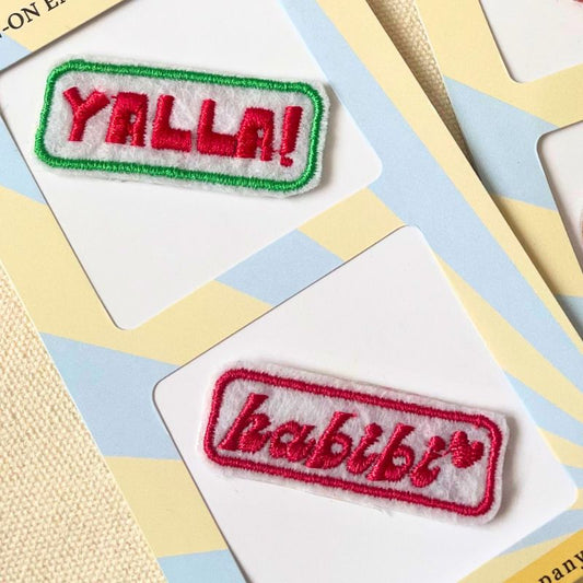 Two embroidered rectangular patches on white backing cards. The top patch reads “YALLA!” in red embroidery with a green border. The second patch is a red-bordered patch reading “HABIBTI” in red on a white background.