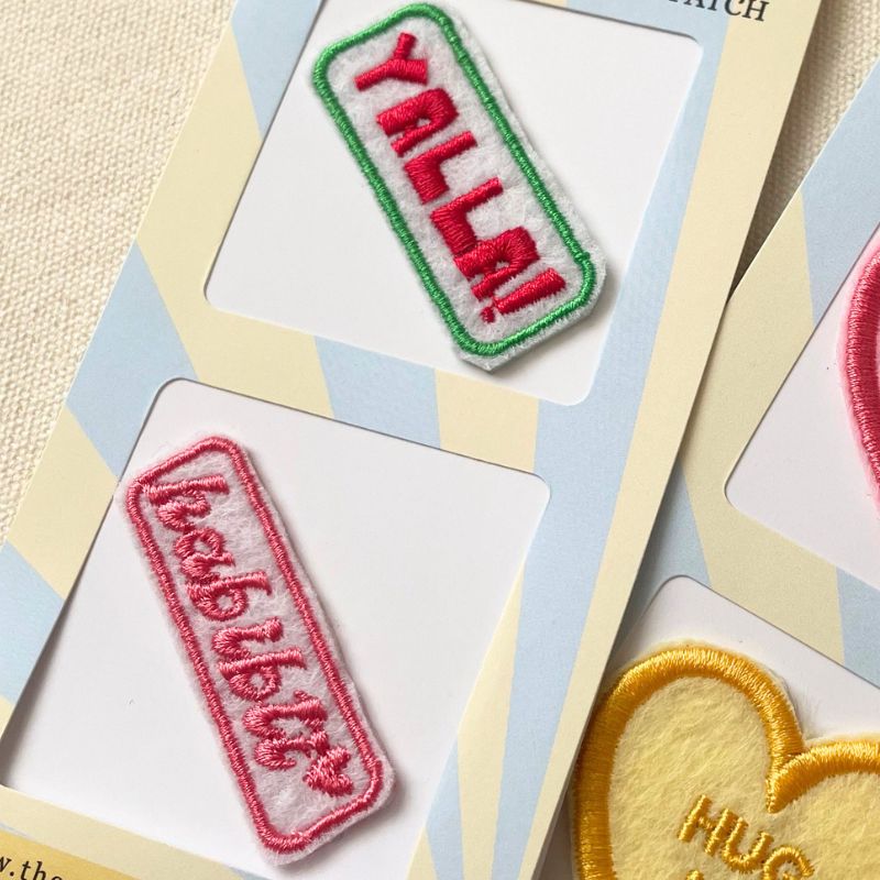 Close-up of embroidered patches displayed on pastel cards. Visible patches include a green-bordered patch reading “YALLA!” in red, a pink-bordered patch reading “HABIBTI” in pink on a white background.